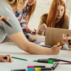 Eine Gruppe von Menschen sitzt um einen Tisch, arbeitet gemeinsam mit Stiften und einem Laptop, hält Notizen fest und diskutiert konzentriert. Verschiedene bunte Stifte und Papier liegen bereit.