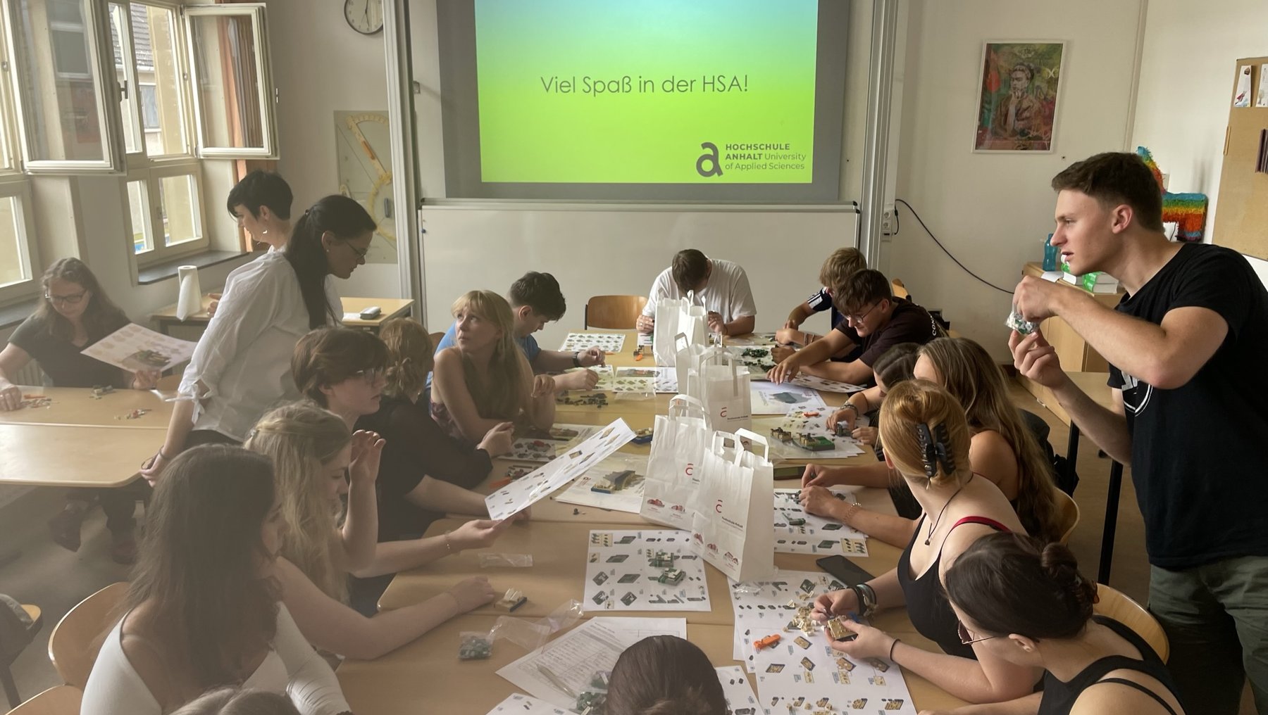 Gruppe von Menschen in einem hellen Raum, die gemeinsam an einem langen Tisch mit großen Arbeitsblättern sitzt, die mit Illustrationen bedruckt sind. Im Hintergrund ist eine Leinwand mit dem Text „Viel Spaß in der HSA!“ zu sehen.