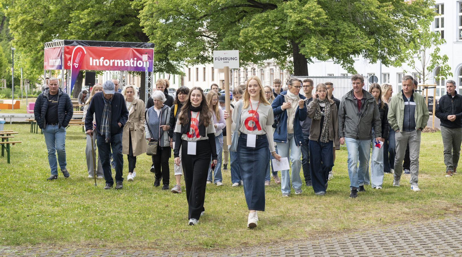 Gruppe von Menschen mit Namensschildern, die auf einem öffentlichen Platz in einer Reihe gehen, vermutlich bei einer Veranstaltung oder Führung. Im Hintergrund ein Infostand mit dem Schild „Information“.