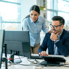 Zwei Personen in einem modernen Büro arbeiten gemeinsam an einem Computer. Eine Person zeigt auf den Bildschirm, während die andere konzentriert zuhört und auf die Tastatur zeigt.