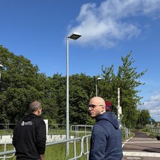 Drei Männer stehen an einer Bahnübergangssperre mit geschlossenen roten und weißen Schranken, im Hintergrund grüne Bäume und ein sonniger Himmel.