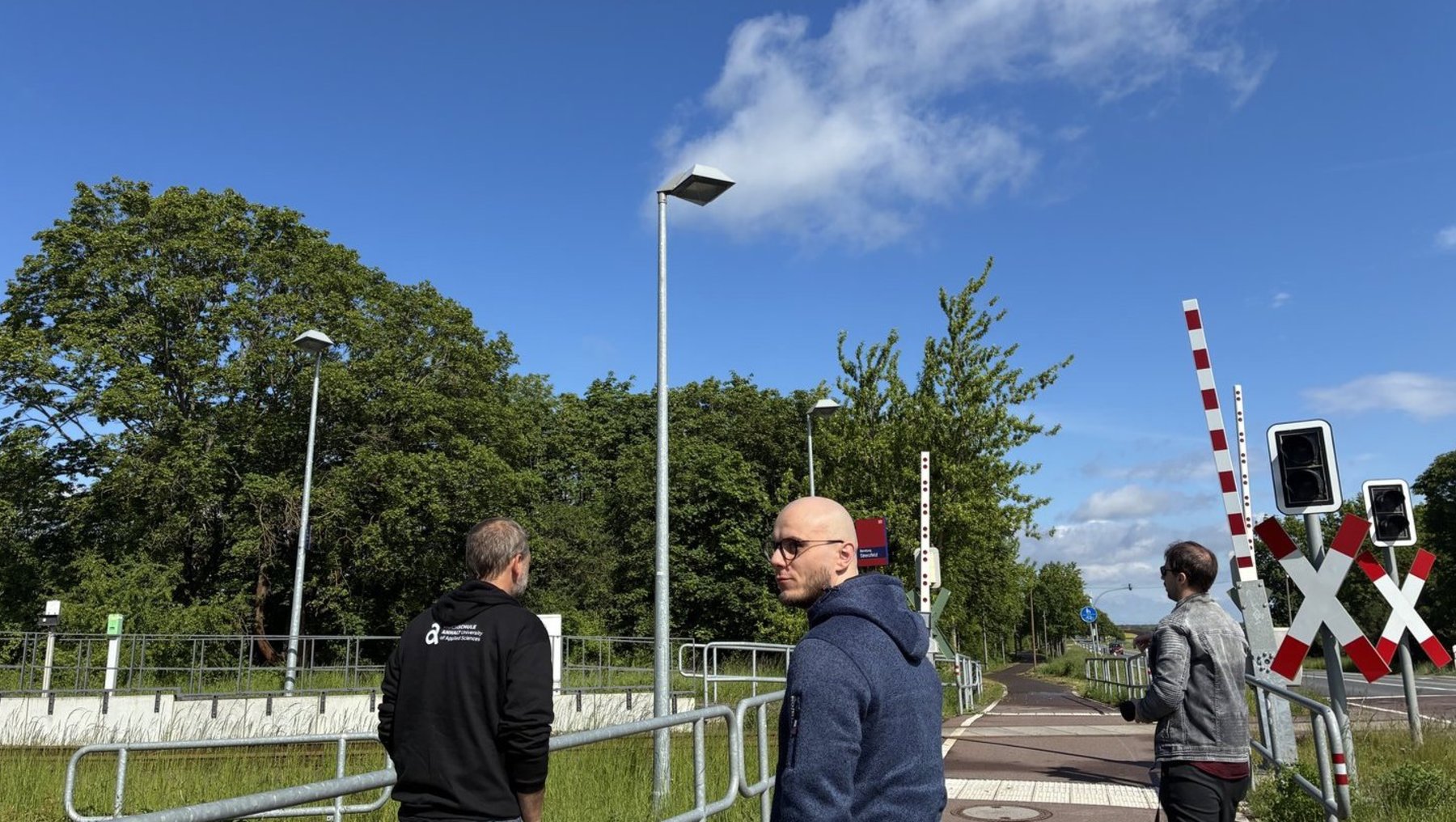 Drei Männer stehen an einer Bahnübergangssperre mit geschlossenen roten und weißen Schranken, im Hintergrund grüne Bäume und ein sonniger Himmel.