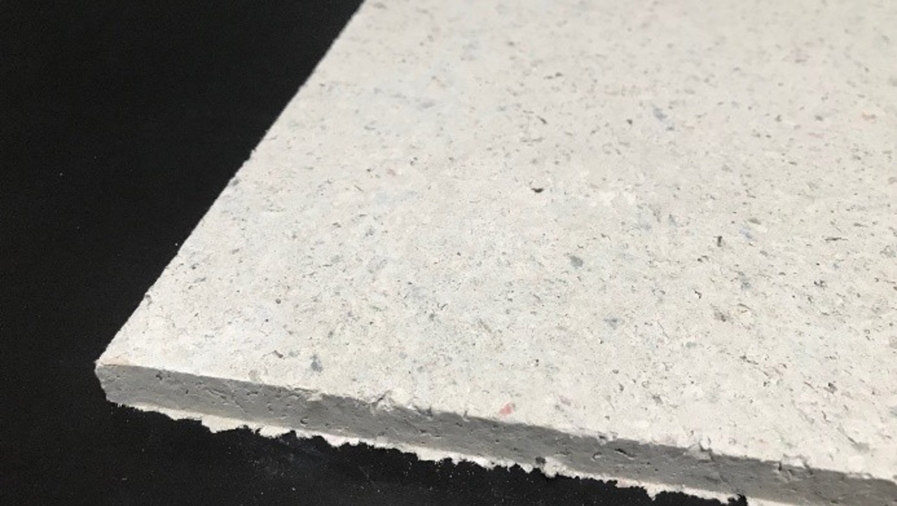Weiße, leicht strukturierte Platte mit rauer Kante, vermutlich aus Beton oder ähnlichem Material, liegt schräg auf dunklem Untergrund. Oberfläche wirkt porös mit kleinen Unebenheiten.