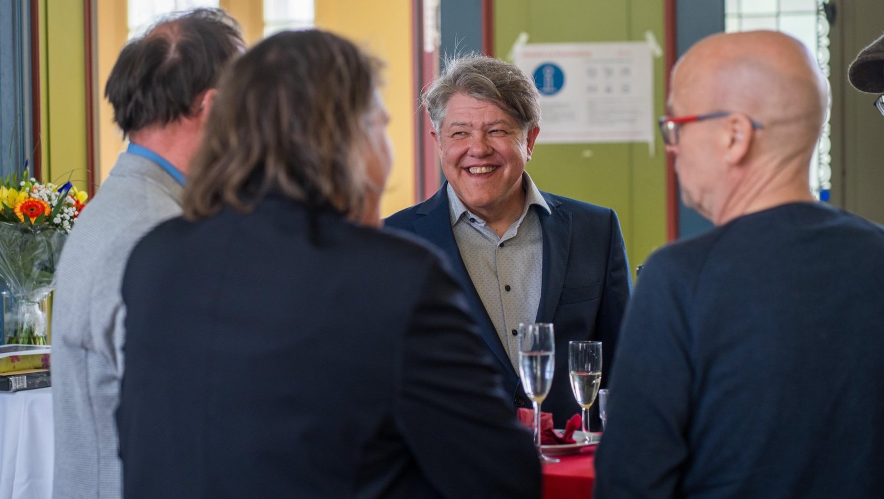 Vier Personen unterhalten sich lächelnd bei einer Veranstaltung, wobei ein Mann im Mittelpunkt mit zwei Gläsern Wein in den Händen steht.