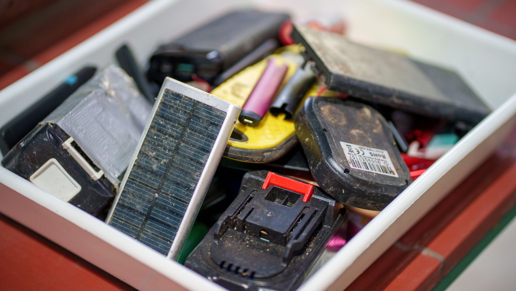 Eine Schublade mit verschiedenen defekten oder alten Batterien, darunter mehrere Akkupacks, ein Ladegerät, ein altes Handy und ein paar Kabel mit Steckverbindungen zeigt Auswahl von möglichen Batterien für das thermische Recycling im Labor der Hochschule Anhalt.