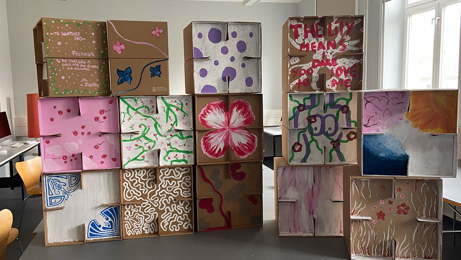 Bunte, kreativ gestaltete Pappkartons in verschiedenen Größen, verziert mit Mustern wie Blumen, geometrischen Formen, Wasserfarben-Effekten und abstrakten Designs, die in einem Raum aufgestellt sind.