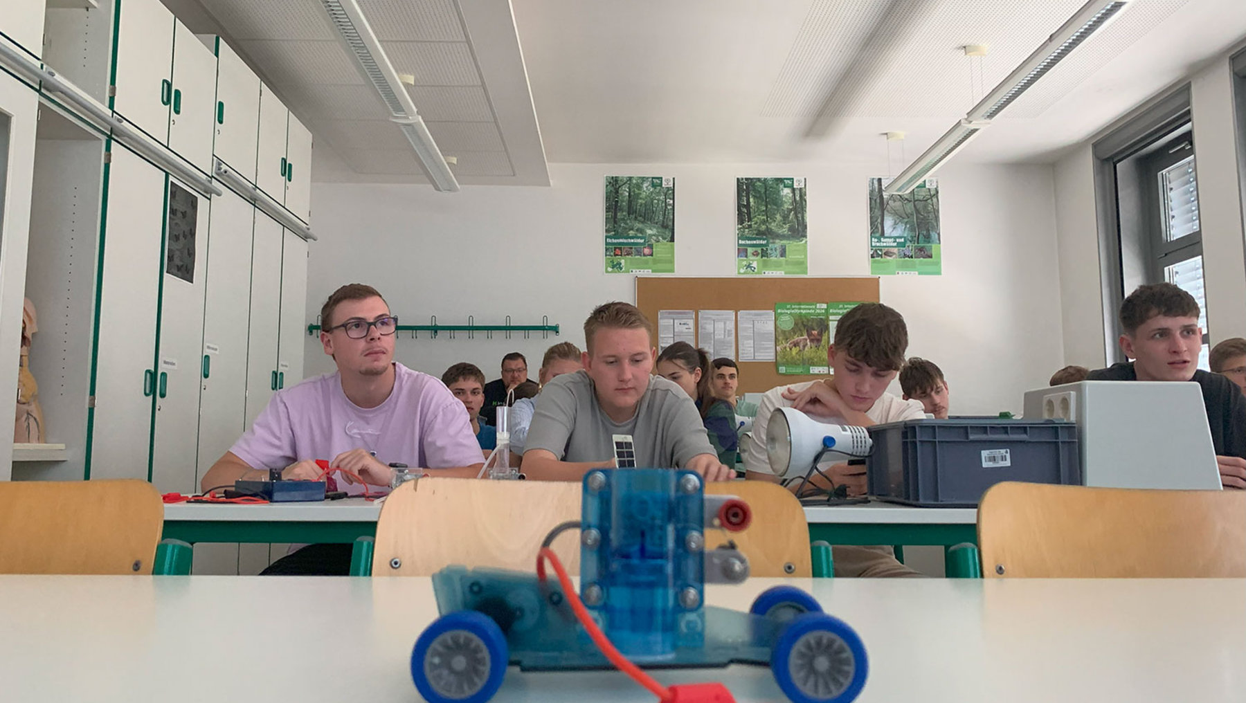 Drei Schüler sitzen in einem Klassenzimmer und arbeiten an einem selbstgebauten, blauen und roten Robotikfahrzeug mit großen blauen Rädern auf einem Tisch. Im Hintergrund weitere Schüler, die konzentriert an Projekten arbeiten.