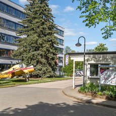 A modern multi-story building with large windows is situated behind a well-maintained lawn and trees. In the foreground, there is a small structure resembling a kiosk or information booth with a poster on it. The scene is bright and sunny with clear skies.