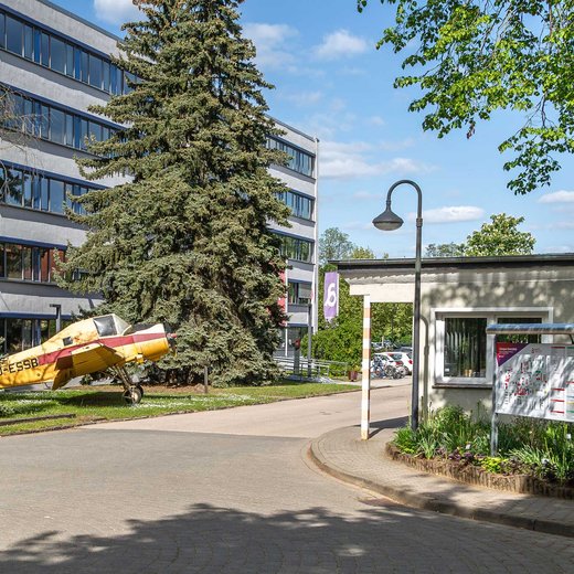 Ein modernes Gebäude mit mehreren Stockwerken steht im Hintergrund. Vor dem Gebäude befindet sich ein großer Baum. Im Vordergrund ist ein kleiner Pavillon mit einer Informationstafel und ein gepflasterter Weg zu sehen. Die Szene wirkt ruhig und ist von Grünflächen umgeben.
