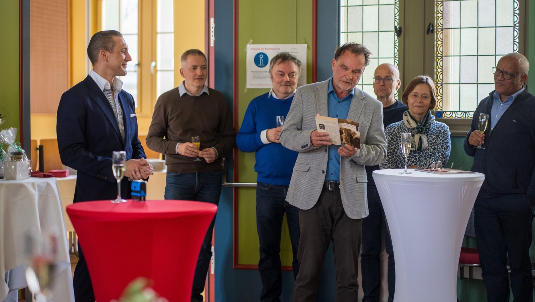 Eine Gruppe von sechs Personen steht in einem Raum und hält eine kleine Feier oder Preisverleihung ab. Ein Mann in der Mitte erhält einen Gegenstand aus den Händen eines anderen, während die anderen zuschauen und einige Gläser in den Händen halten.