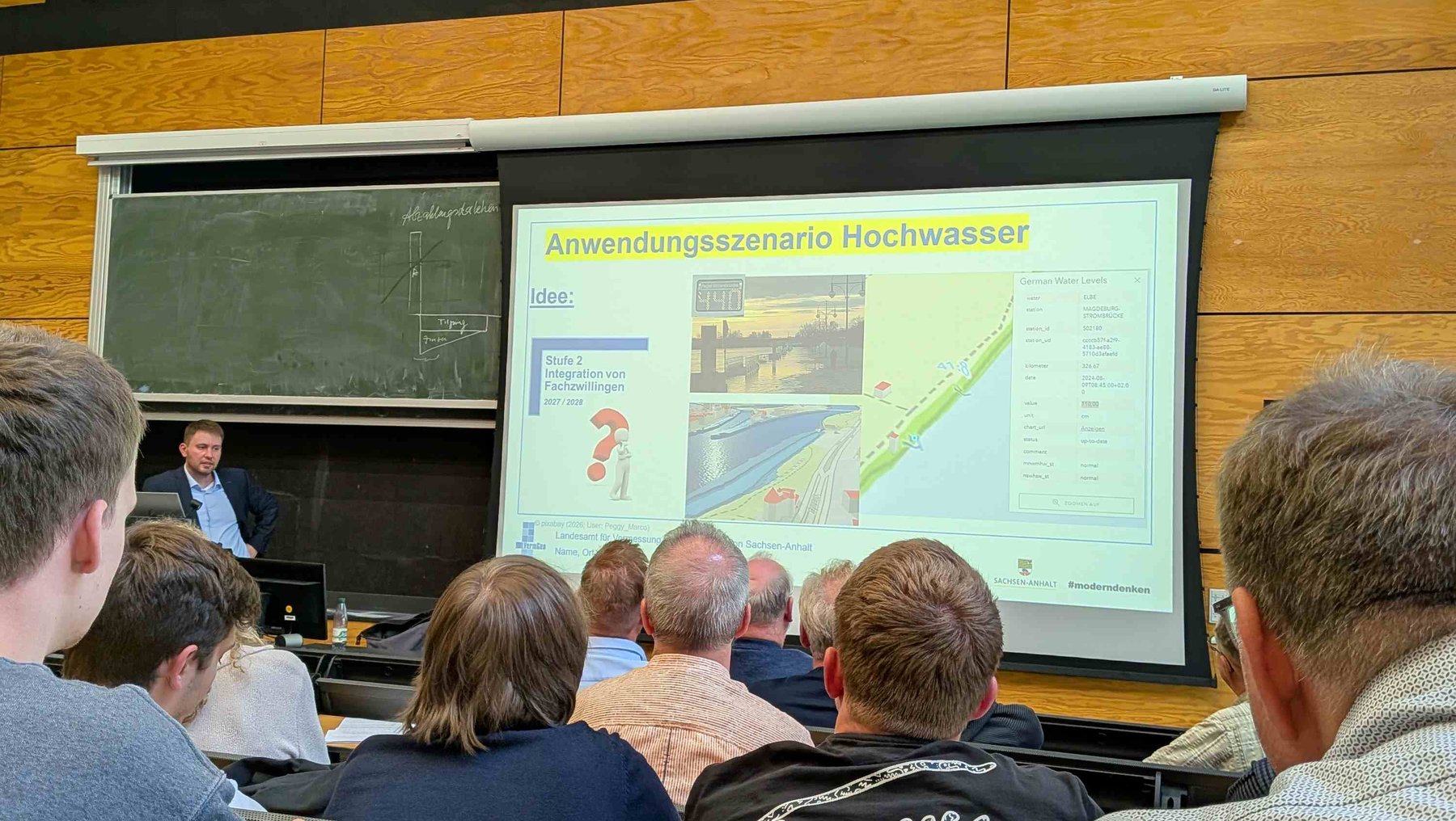 Ein Vortragsraum mit Publikum, das einer Präsentation zu Hochwasserszenarien und Anwendungsbeispielen folgt. Auf dem Bildschirm sind Diagramme und Karten mit Überschwemmungsgebieten sowie ein Foto einer überschwemmten Straße zu sehen. Ein Referent steht am Podium und spricht zum Thema.