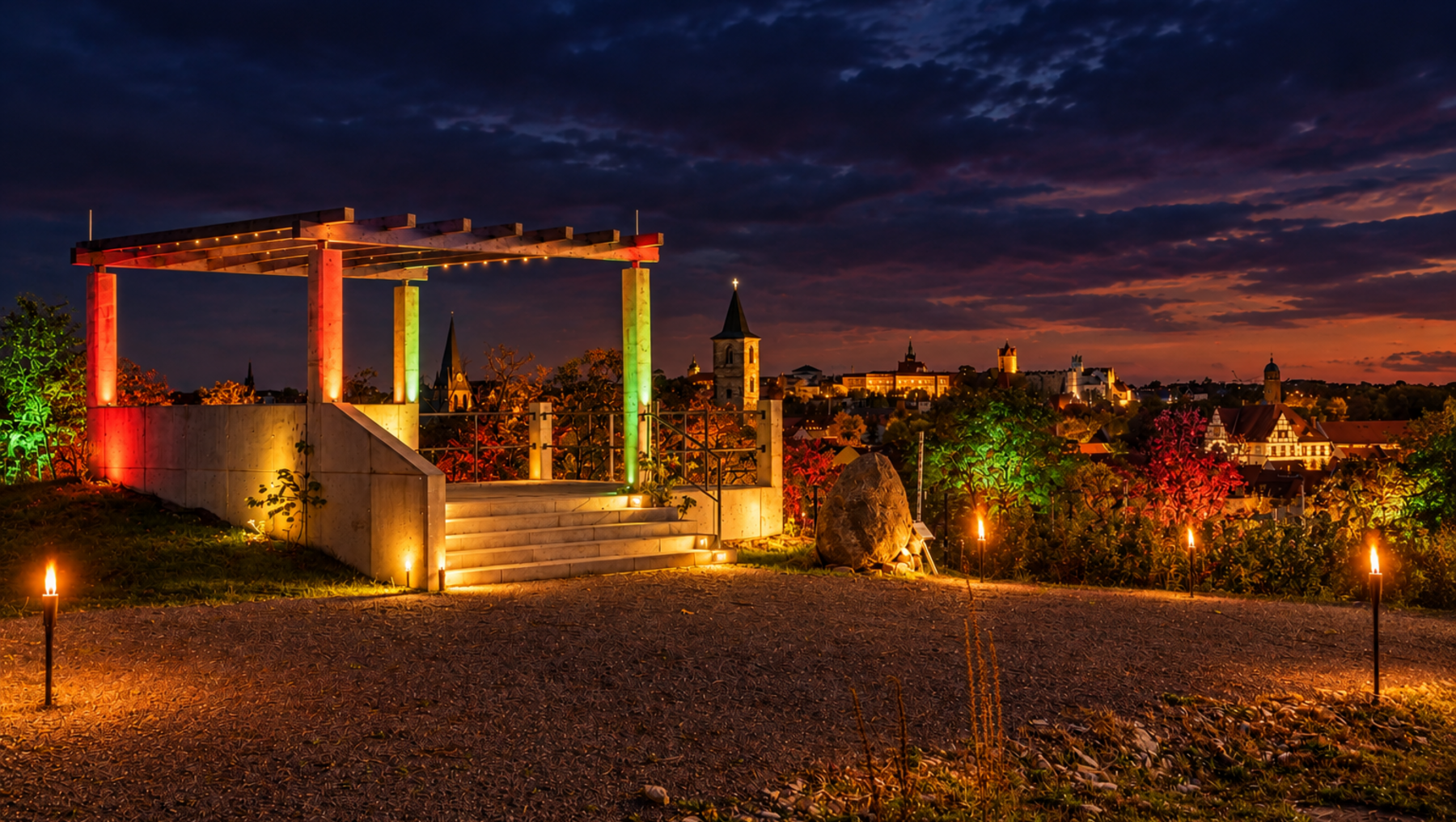Abendliche Aussicht auf eine beleuchtete Aussichtsplattform mit Treppe, umgeben von buntem Herbstlaub. Im Hintergrund eine Stadt mit Kirchturm und beleuchteten Gebäuden bei Dämmerung.