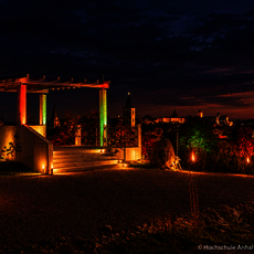 Abendliche Aussicht auf eine beleuchtete Aussichtsplattform mit Treppe, umgeben von buntem Herbstlaub. Im Hintergrund eine Stadt mit Kirchturm und beleuchteten Gebäuden bei Dämmerung.