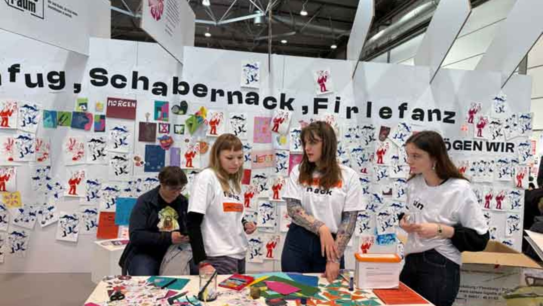 Vier Personen stehen an einem Infotisch mit dem Titel „Fug, Schabernack, Firlefanz“ und halten bunte, kreative Materialien wie Puzzle- und Bastelteile. Im Hintergrund hängen Illustrationen von Menschen in verschiedenen Posen.