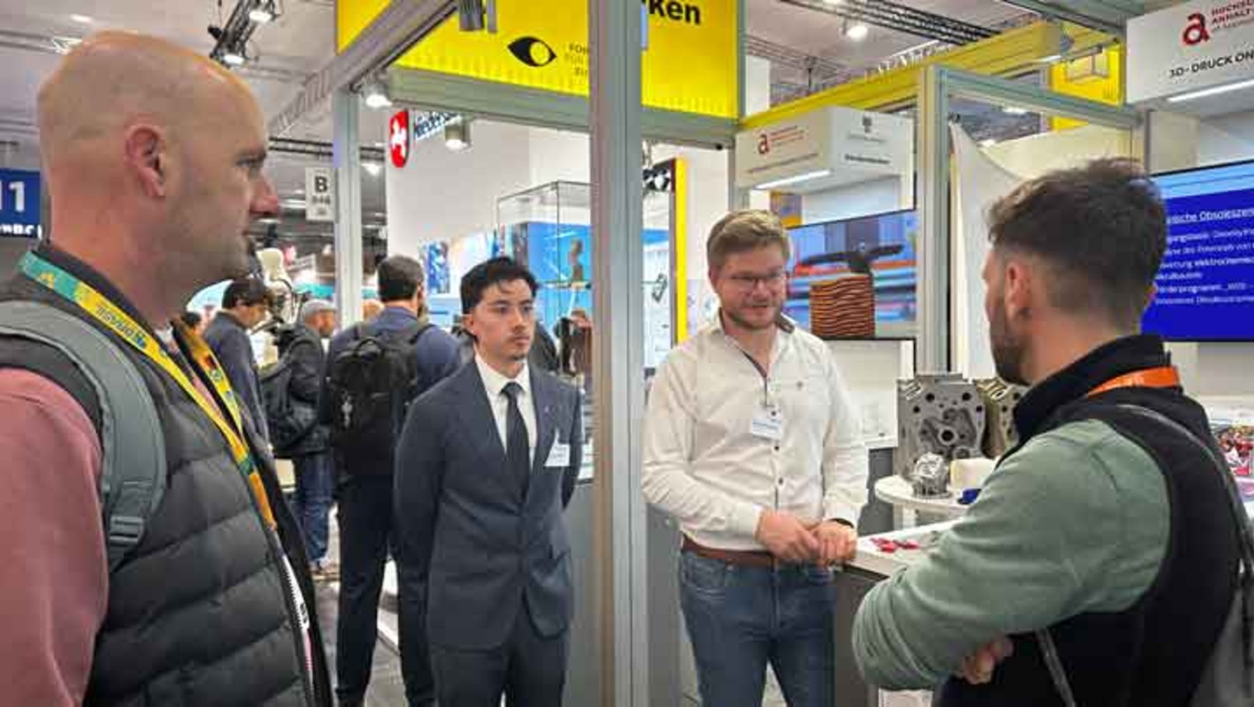 Vier Männer auf einer Messe: Einer im Anzug, zwei in Business-Casual und einer mit Rucksack, die sich an einem Messestand mit Technikausstellungen unterhalten und sich die Hand reichen.