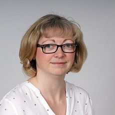 A woman with short blonde hair and glasses is looking slightly to the side. She is wearing a white blouse with small black dots. The background is plain and light-colored.
