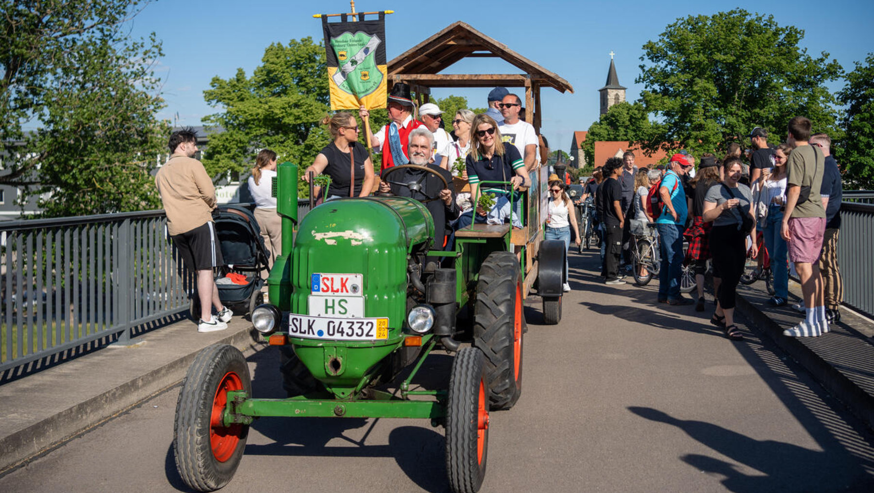 Grüner Traktor mit Kennzeichen „SLK 04332“ fährt über eine Brücke, auf dem Dach eine Flagge mit Aufschrift „HS“. Mehrere Personen sitzen darauf, im Hintergrund stehen Zuschauer an der Brüstung.