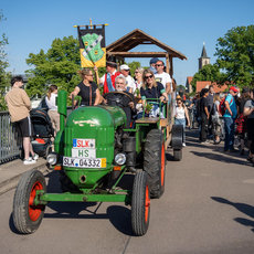 Grüner Traktor mit Kennzeichen „SLK 04332“ fährt über eine Brücke, auf dem Dach eine Flagge mit Aufschrift „HS“. Mehrere Personen sitzen darauf, im Hintergrund stehen Zuschauer an der Brüstung.