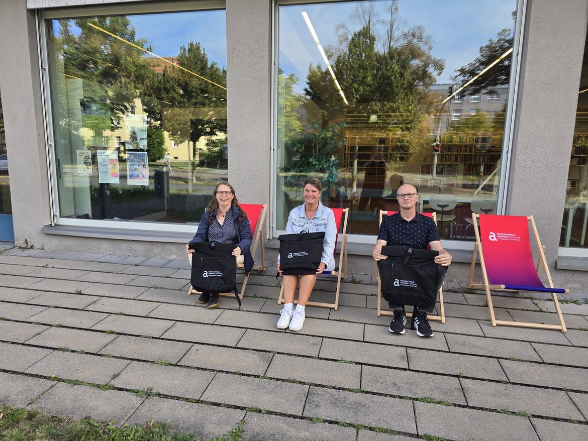 Drei Personen sitzen auf Klappstühlen vor einem Gebäude mit großen Fenstern. Zwei Frauen und ein Mann lächeln in Richtung Kamera. Hinter ihnen ist ein bunter Werbeaufsteller mit Logo und Text zu sehen.