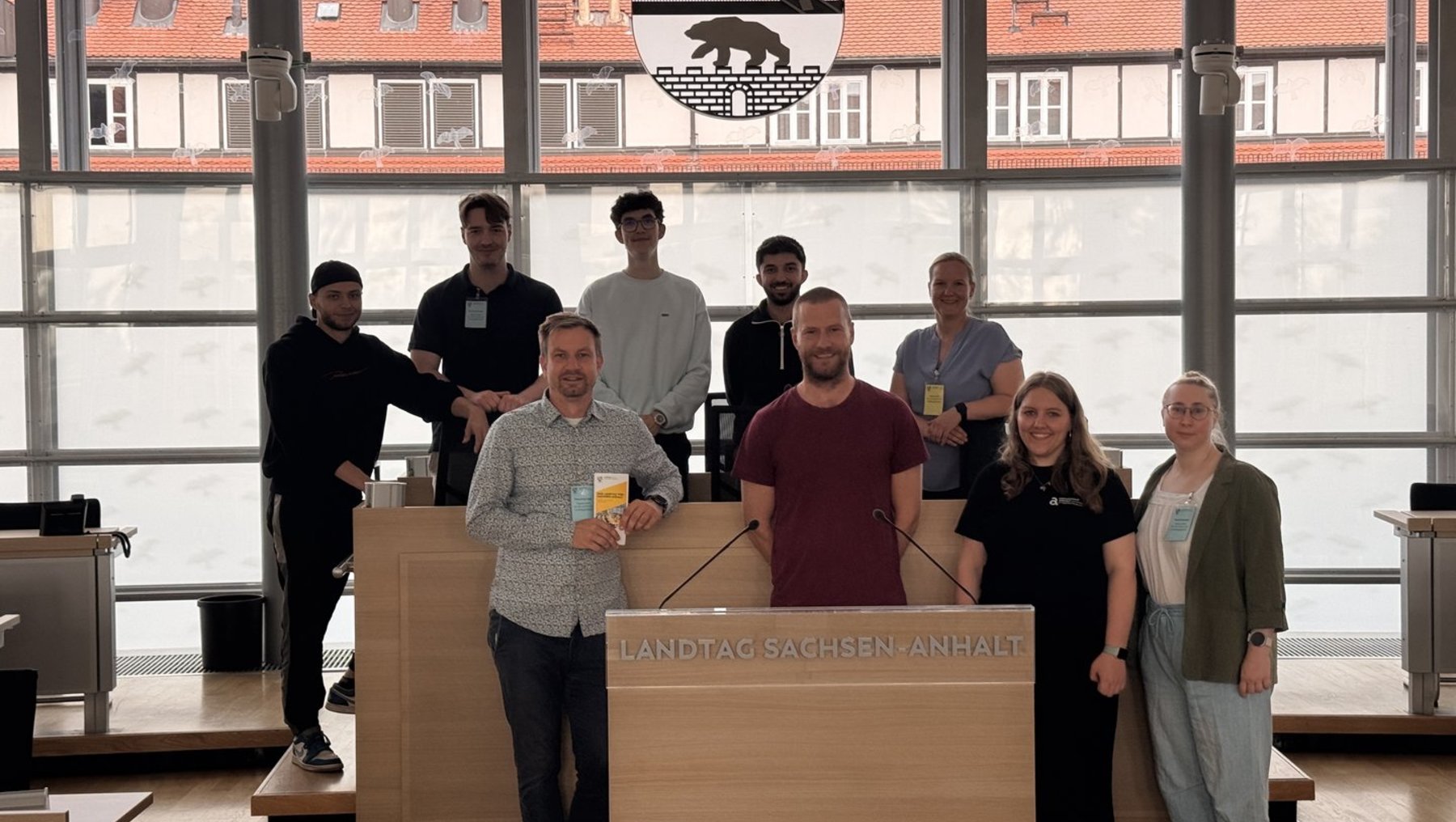 Gruppe von zwölf Personen steht vor einem Podium mit der Aufschrift „Landtag Sachsen-Anhalt“ und hält kleine Becher. Sie wirken zufrieden und posieren für ein Gruppenfoto.