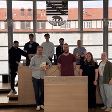 Gruppe von zwölf Personen steht vor einem Podium mit der Aufschrift „Landtag Sachsen-Anhalt“ und hält kleine Becher. Sie wirken zufrieden und posieren für ein Gruppenfoto.