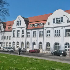 A large, white, multi-story building with a red-tiled roof and numerous arched windows. The building has a symmetrical design with a central section featuring a clock. Several cars are parked in front of the building, and a grassy area with scattered flowers is visible in the foreground.