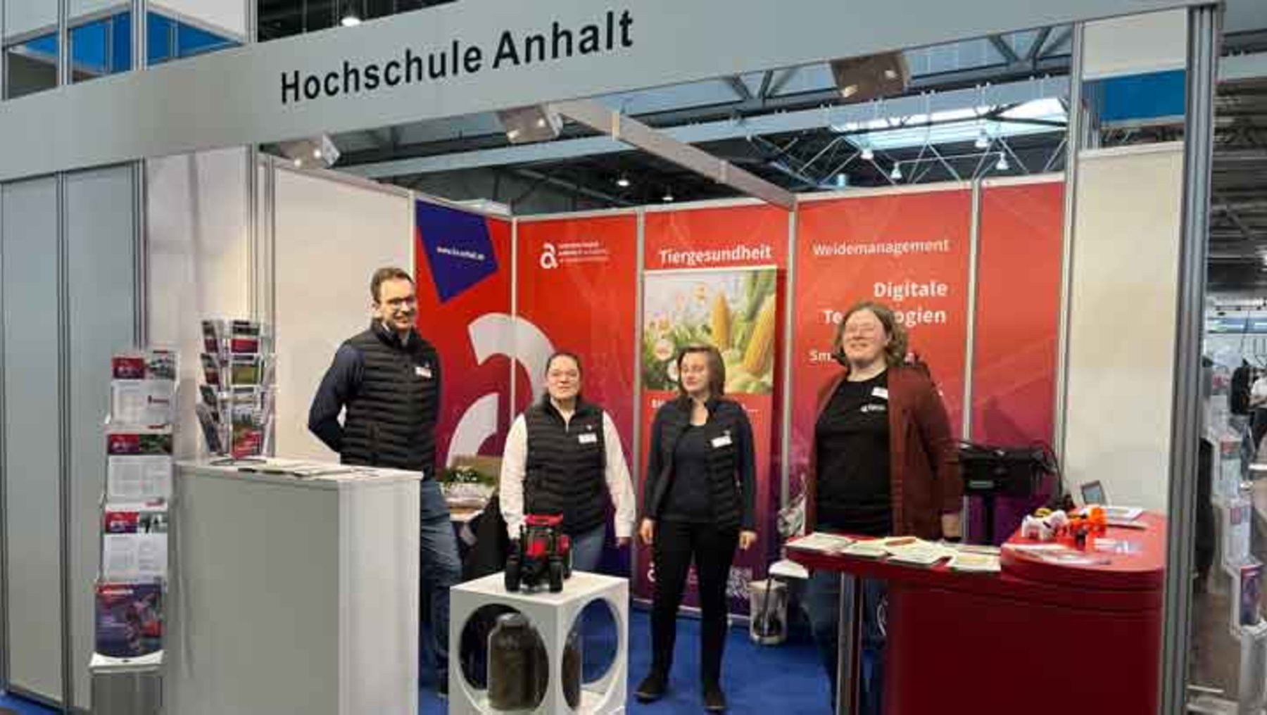 Drei Personen stehen lächelnd hinter einem Messestand der Hochschule Anhalt mit dem Logo und den Themen Terpenidentität, digitale Transformation und Weidemanagement. Im Hintergrund sind Infomaterialien und ein Tisch mit Prospekten zu sehen.