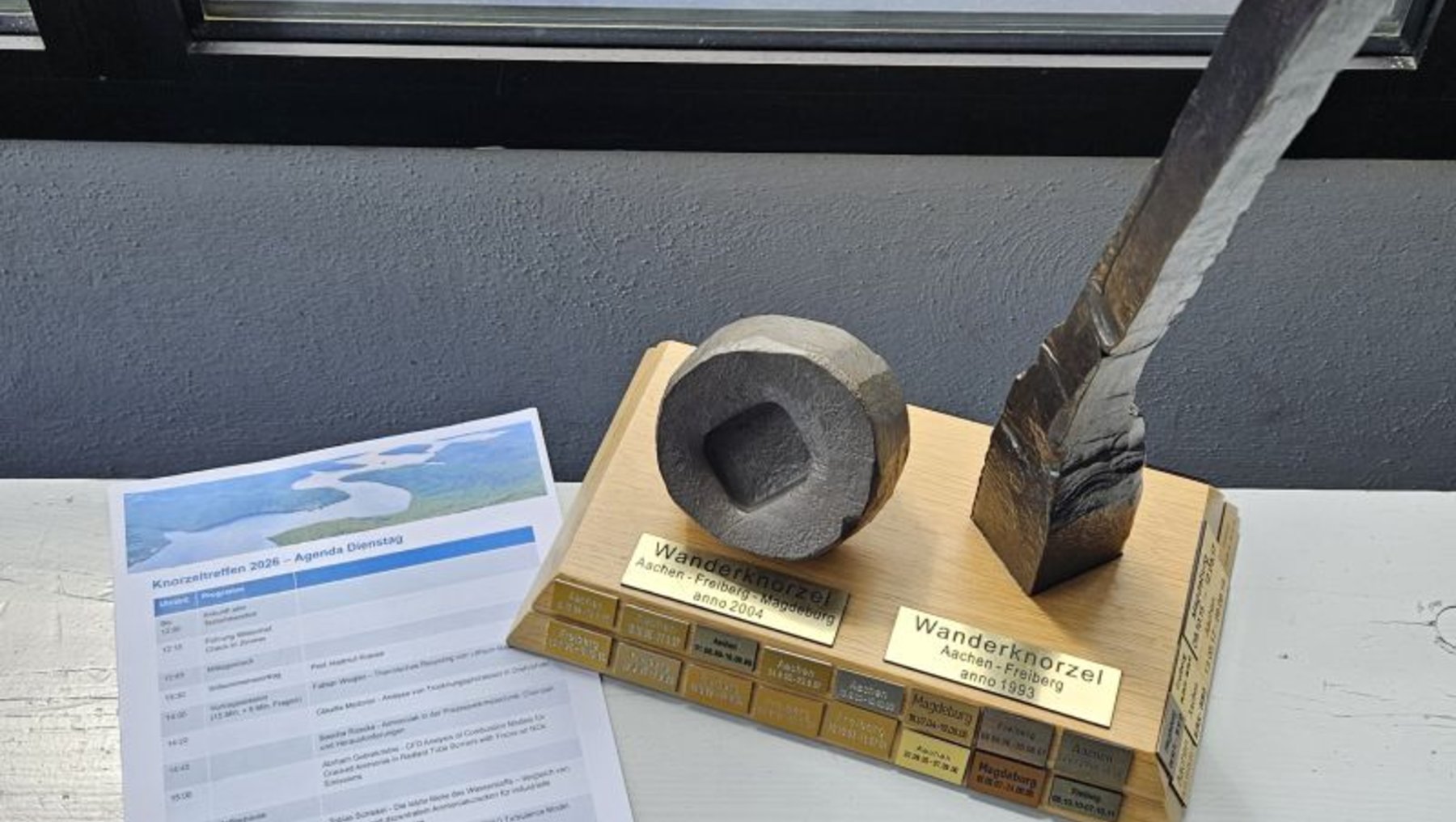 Auf einem Tisch liegt ein altmodischer Füllfederhalter mit einem Tintenfass auf einem Holzsockel. Daneben befindet sich ein Dokument mit Formularen und einem blauen Logo.