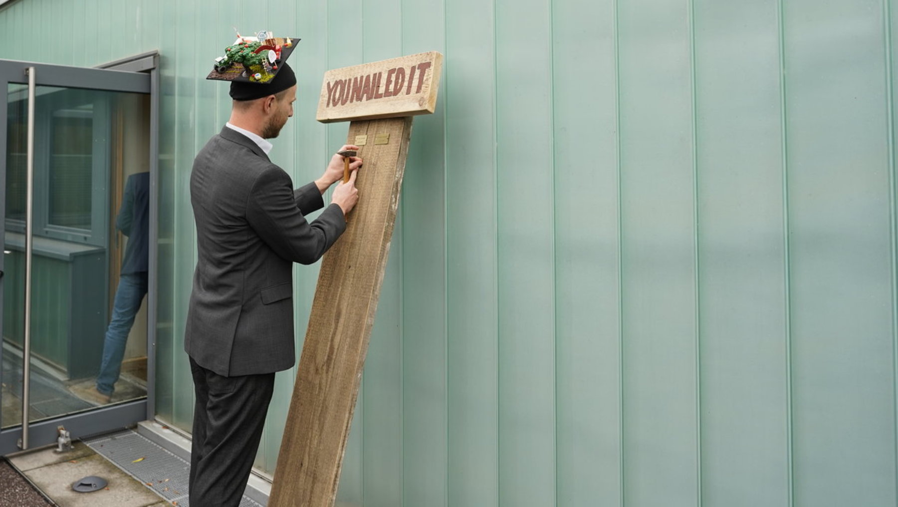 Ein Mann in Anzug befestigt eine Holztafel mit der Aufschrift „You nailed it“ an einer grünen Wand. Im Hintergrund stehen mehrere Personen, die zuschauen.