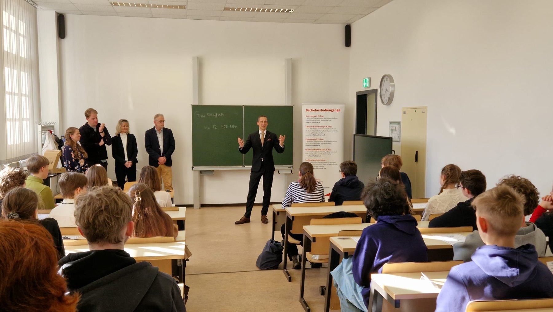 Ein Mann steht vor einer Klasse aus Schülern im Klassenzimmer und hält eine Rede oder Präsentation, während mehrere Personen im Hintergrund stehen und zuhören.
