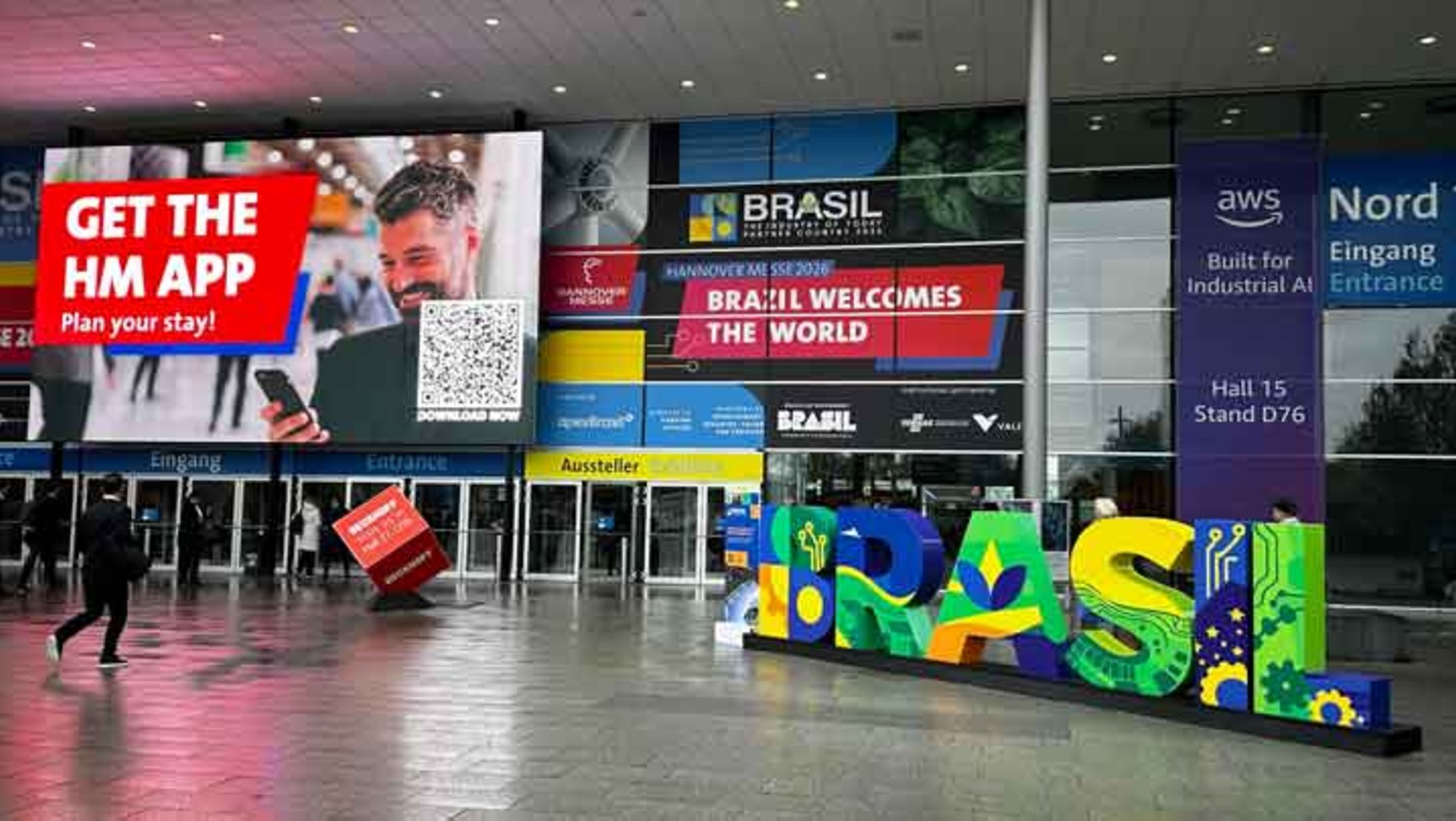 Großes, buntes Schild mit dem Schriftzug „BRASIL“ in großen, farbigen Buchstaben am Eingang einer Messe. Dahinter Banner mit „BRASIL WELCOMES THE WORLD“ und Hinweisen zu Standorten.