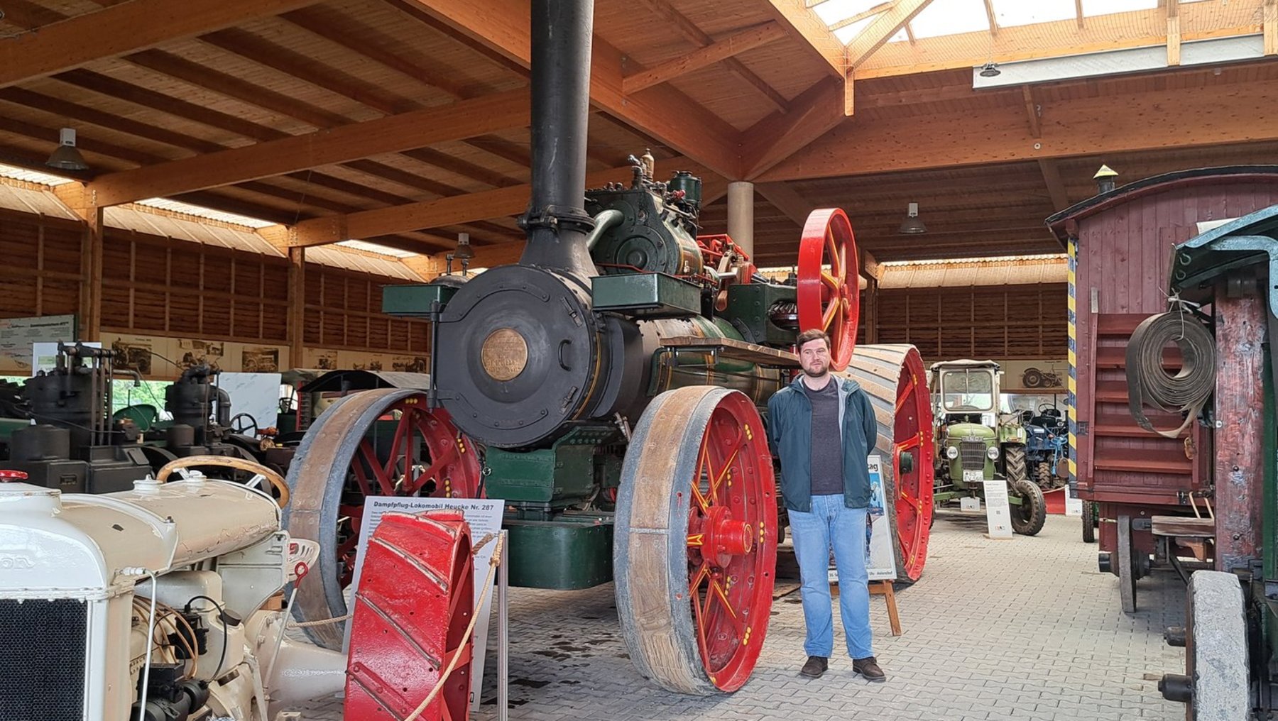 Historische Dampfmaschine mit großen roten Rädern und grünem Kessel in einer Halle, daneben ein Mann in blauer Jacke und Jeans, der vor den Maschinen steht.