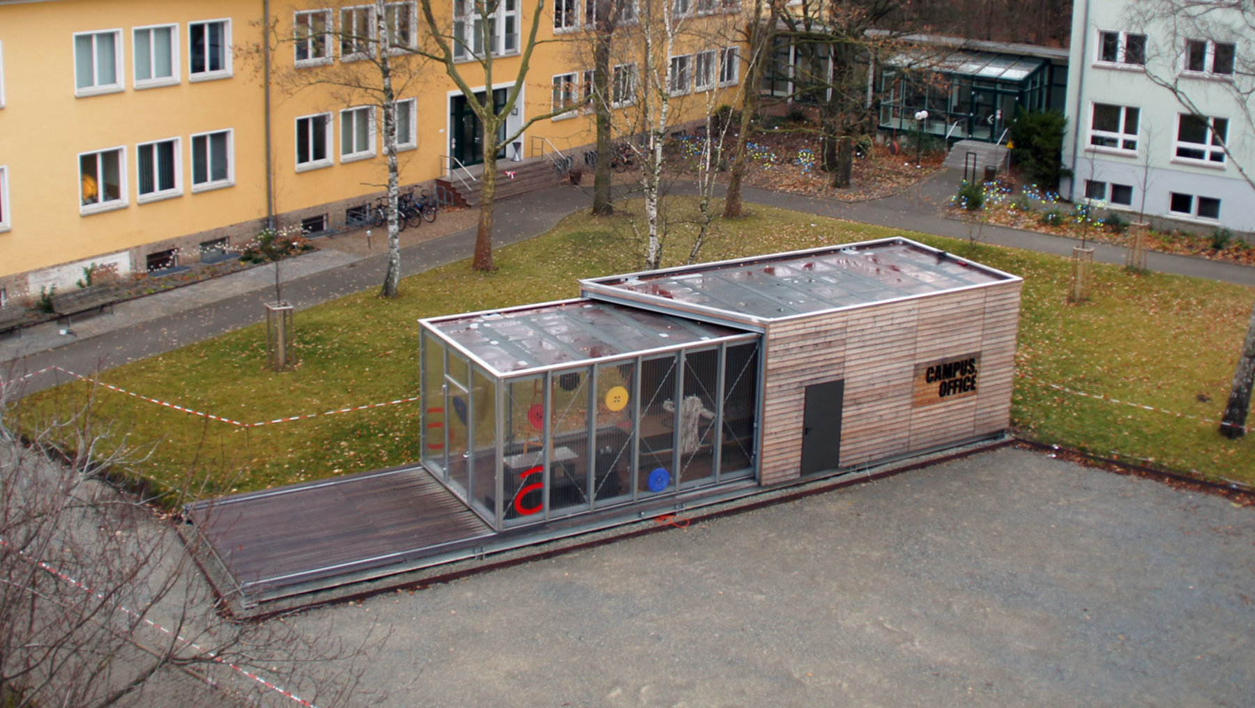 Ein kleines, modernes Gebäude mit Glasfronten und einem Holzrahmen steht auf einem Betonweg. Es ist von einem Rasen und Bäumen umgeben, mit Wohnhäusern im Hintergrund. Das Gebäude scheint als Ausstellungsraum oder kleines Café genutzt zu werden.