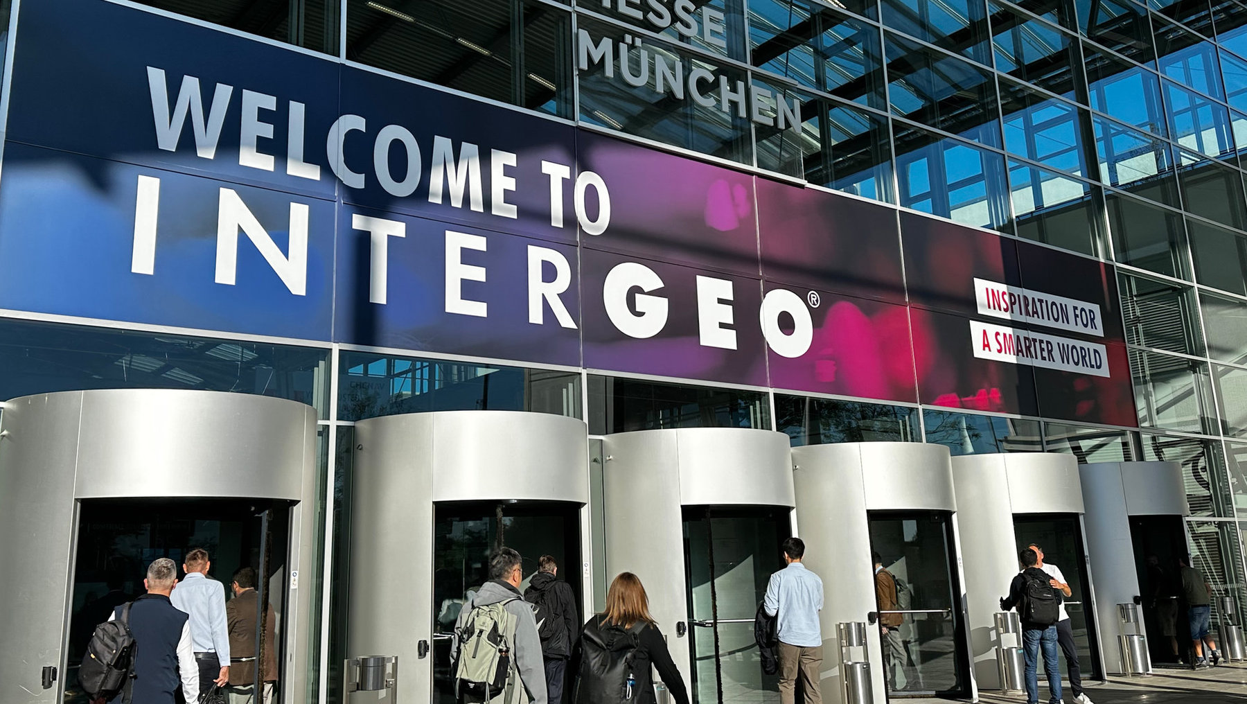 Willkommensbereich der INTERGEO mit großen Buchstaben auf Glasfront, davor Menschen, die den Eingang betreten. Veranstaltungshinweis mit dem Slogan Inspiration for a Smarter World.