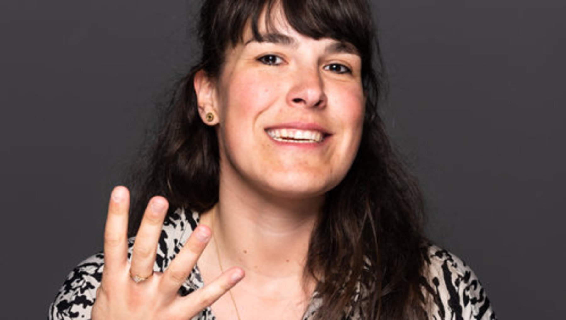 Eine lächelnde Person mit langen Haaren winkt mit der Hand, an der drei Finger gespreizt sind, während sie ein gemustertes Oberteil trägt.
