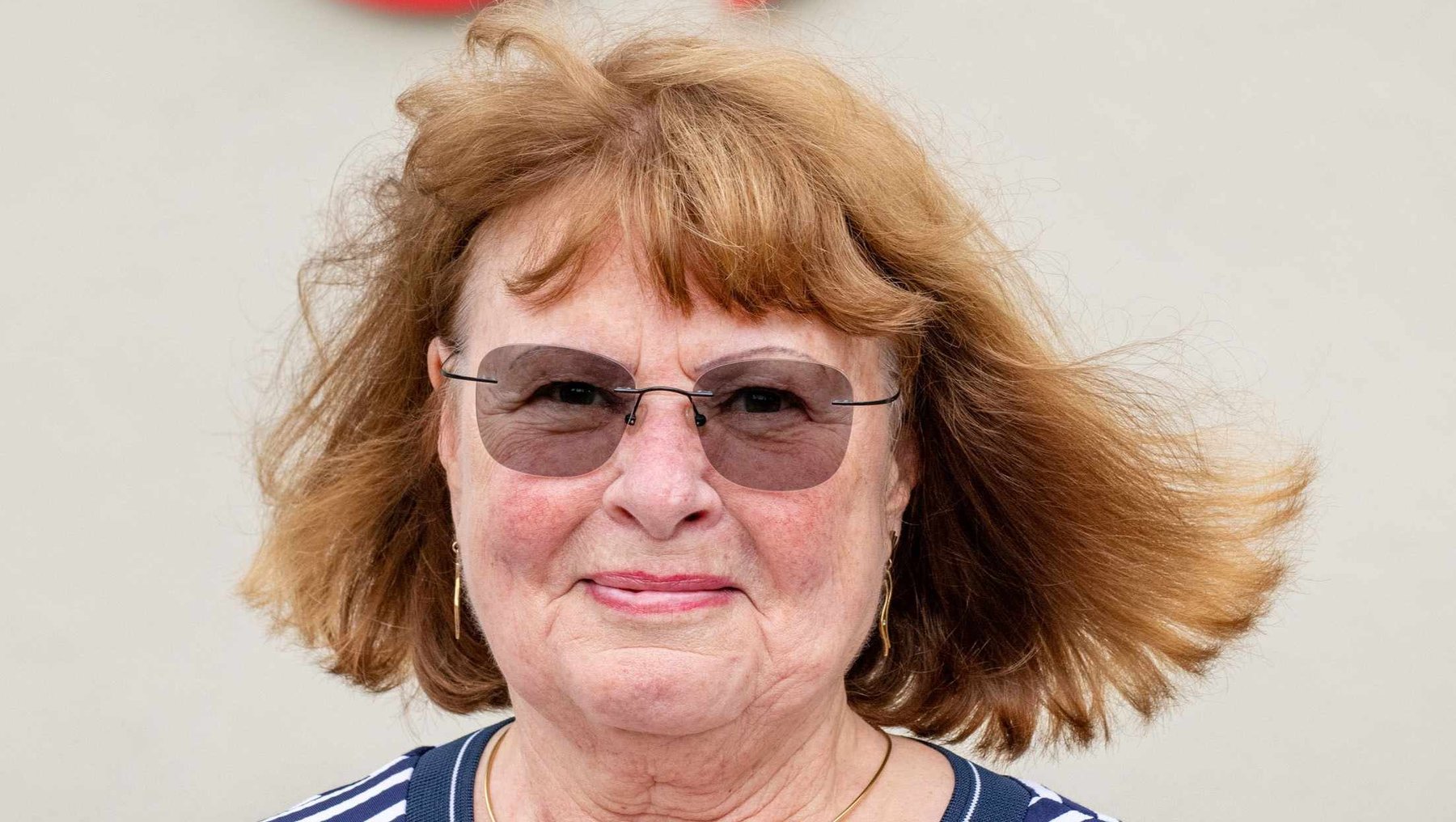 Eine lächelnde Frau mit kurz geschnittenen, hellblonden Haaren und Brille, die ein blau-weiß gestreiftes V-Ausschnitt-Shirt trägt. Im Hintergrund ein Gebäude mit dem Logo der Hochschule Anhalt.