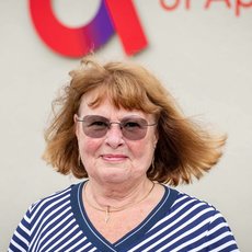 Eine lächelnde Frau mit kurz geschnittenen, hellblonden Haaren und Brille, die ein blau-weiß gestreiftes V-Ausschnitt-Shirt trägt. Im Hintergrund ein Gebäude mit dem Logo der Hochschule Anhalt.