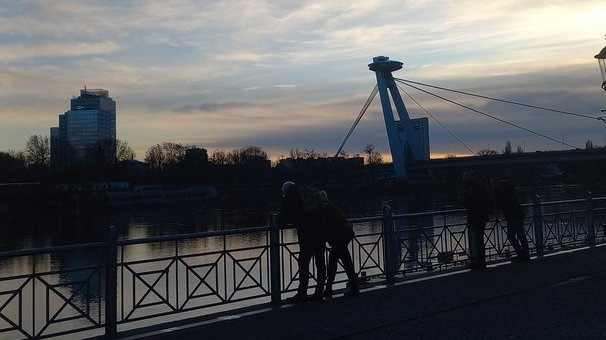 Zwei Personen spazieren bei Sonnenuntergang über eine Brücke mit Geländer, im Hintergrund ein Kran und ein Hochhaus am Himmel mit bläulich-orangefarbenen Wolken.