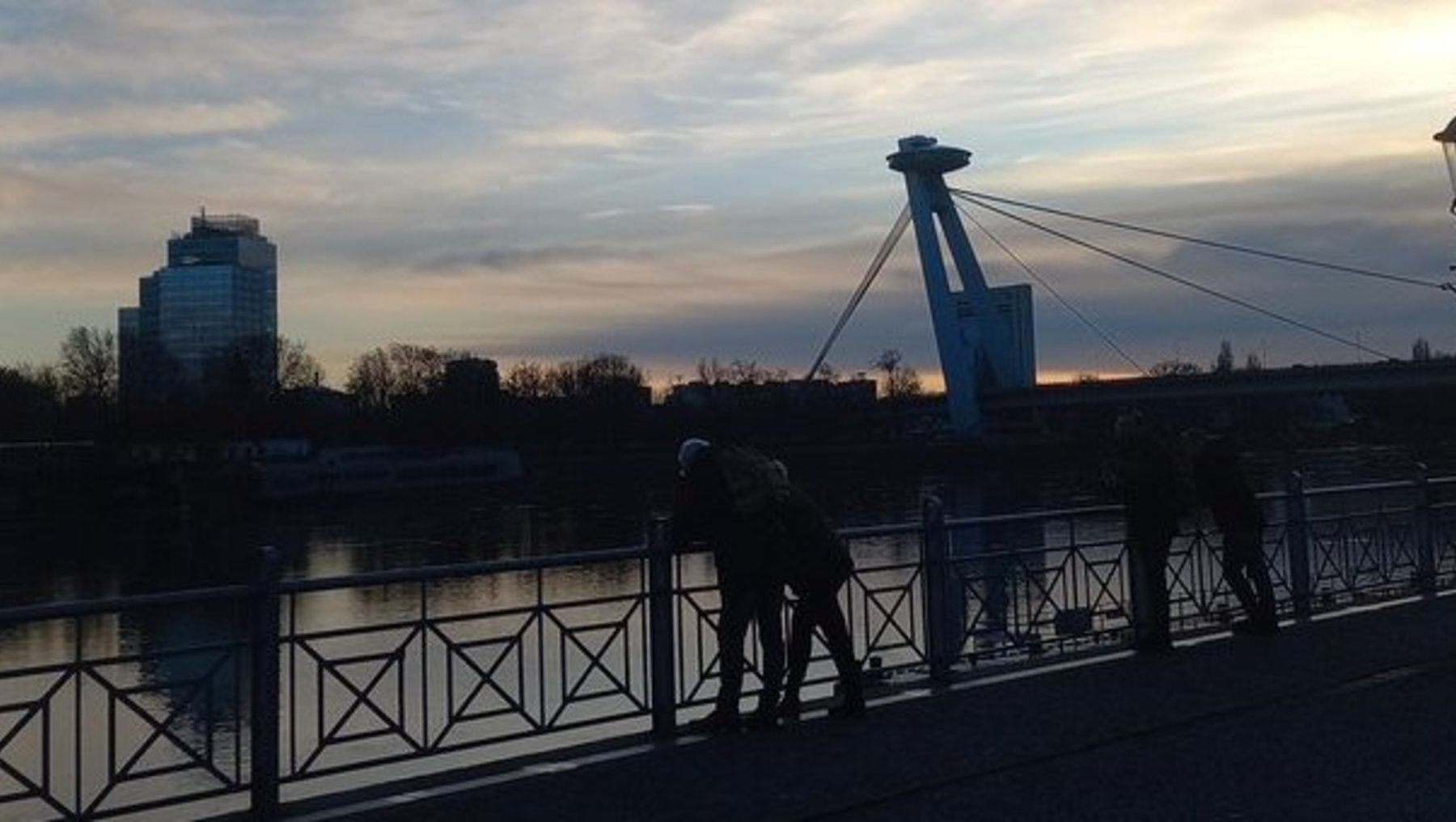 Zwei Personen spazieren bei Sonnenuntergang über eine Brücke mit Geländer, im Hintergrund ein Kran und ein Hochhaus am Himmel mit bläulich-orangefarbenen Wolken.