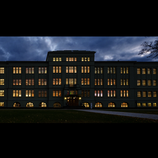 Großes, symmetrisch aufgebautes Gebäude mit klassischer Architektur, beleuchtet in der Abenddämmerung oder Nacht, mehrere Fensterreihen und zentraler Eingang mit Treppe.