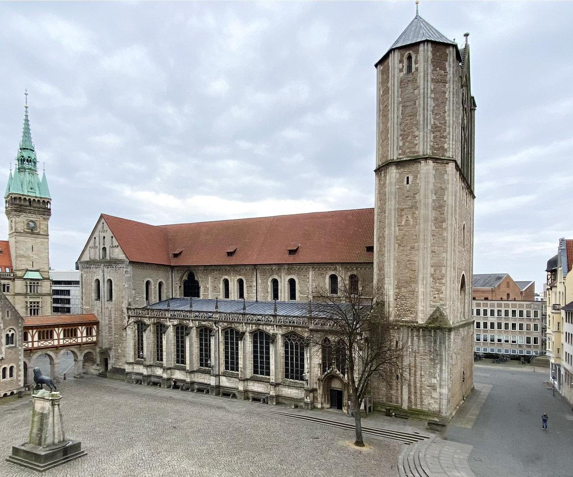 Großes historisches Kirchengebäude aus Stein mit rotem Dach, bestehend aus einem hohen Turm auf der rechten Seite und einem längeren Hauptschiff mit Arkaden. Im Hintergrund weitere gotische Architektur mit spitzem Turm. Vor dem Gebäude ein leerer Platz mit wenigen Bäumen und einer Statue.
