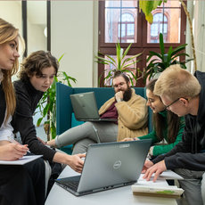 Vier Personen sitzen in einem modernen Büro oder Lernraum auf einer Couch und arbeiten gemeinsam an einem Laptop, während eine Person Notizen macht.