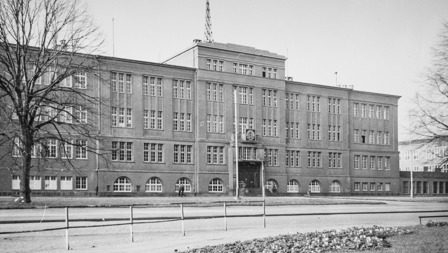 Historisches, mehrstöckiges Schulgebäude mit zentralem Haupteingang, symmetrisch angeordneten Fenstern und einem markanten Turmaufsatz, umgeben von kahlen Bäumen und einem gepflegten Vorplatz.