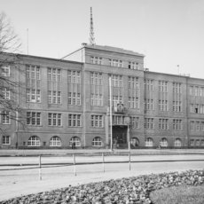 Historisches, mehrstöckiges Schulgebäude mit zentralem Haupteingang, symmetrisch angeordneten Fenstern und einem markanten Turmaufsatz, umgeben von kahlen Bäumen und einem gepflegten Vorplatz.