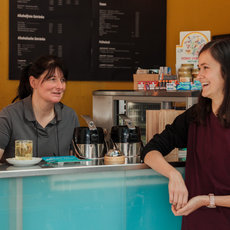 Two women are at a coffee shop counter. One woman, behind the counter, is holding a pen and smiling. The other woman, in front of the counter, is also smiling and leaning slightly forward. The counter has a coffee machine and a glass of water.