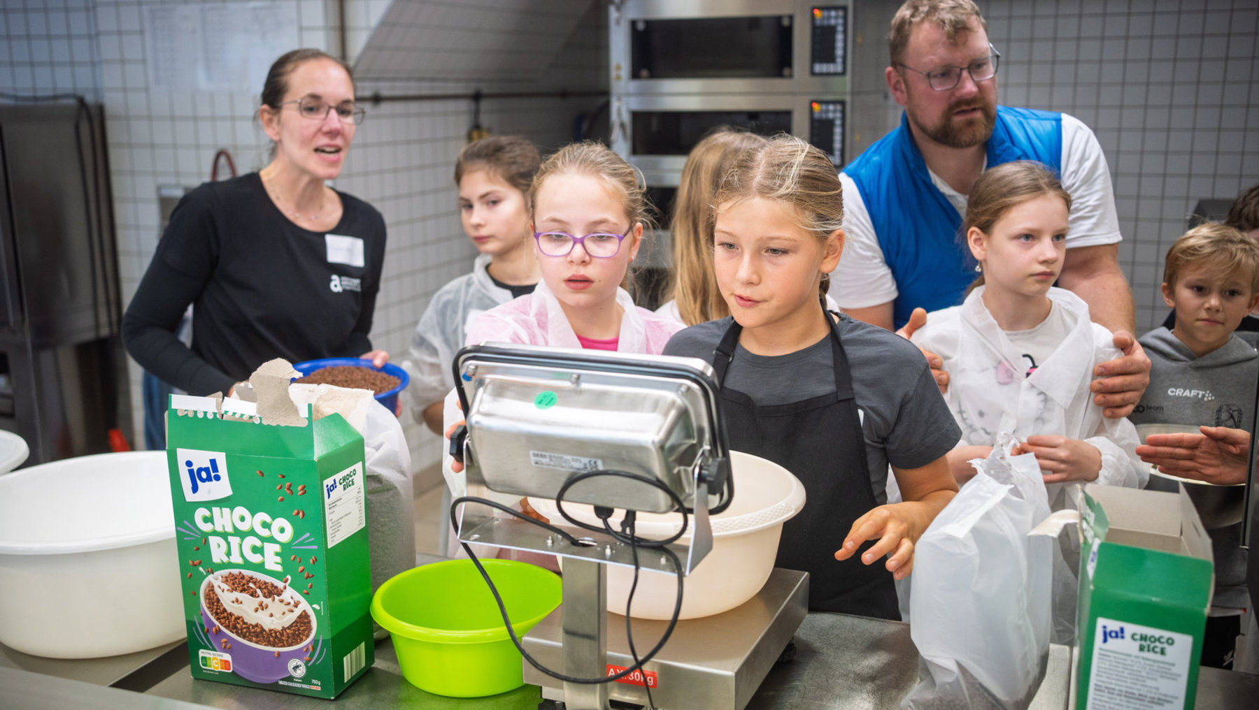 Eine Gruppe von Kindern und zwei Betreuer in einer Küche, die gemeinsam Schokoladenreis zubereiten. Auf dem Tisch stehen Packungen mit Schoko-Reis-Mischung, eine Küchenwaage und ein Mixgerät. Die Kinder beobachten aufmerksam den Prozess.
