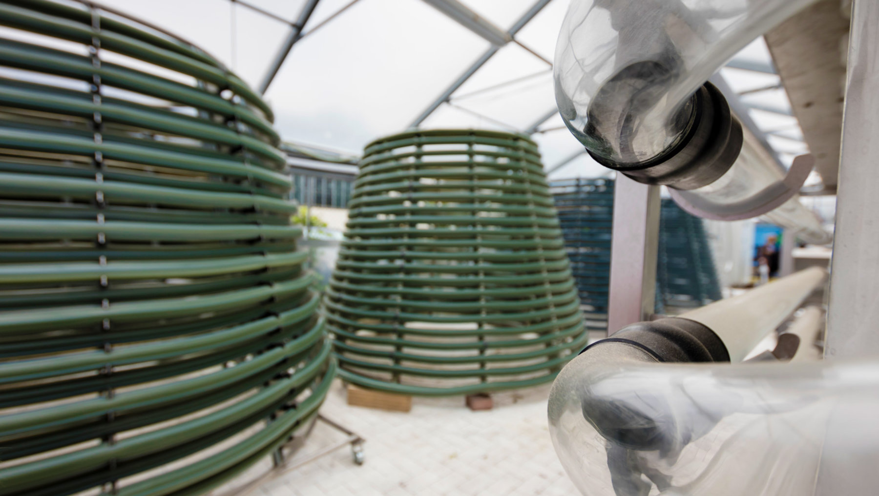 The image shows stacks of coiled, green plastic vertical planters, likely for hydroponic or indoor gardening use in a greenhouse setting. Several these containers are placed neatly on shelves.