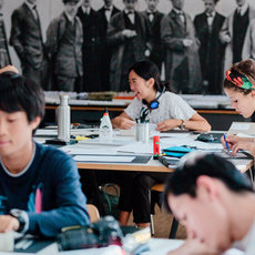A group of people are seated at tables in a classroom setting, working on various tasks. They appear to be focused on their work, with papers, notebooks, and writing instruments in front of them. The background features a large black-and-white photograph of a group of people.