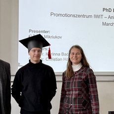 Drei Personen stehen vor einer Präsentationstafel zur PhD-Verteidigung an der Anhalt University of Applied Sciences am 16. März 2026. Der Mittelste trägt eine Doktorhut und ist als Präsentator Evgeny Mikriukov genannt. Links und rechts stehen zwei Gutachter, eine Frau und ein Mann.