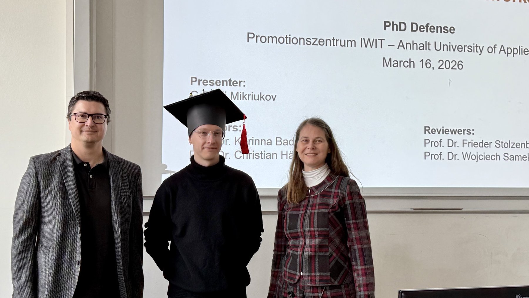 Drei Personen stehen vor einer Präsentationstafel zur PhD-Verteidigung an der Anhalt University of Applied Sciences am 16. März 2026. Der Mittelste trägt eine Doktorhut und ist als Präsentator Evgeny Mikriukov genannt. Links und rechts stehen zwei Gutachter, eine Frau und ein Mann.